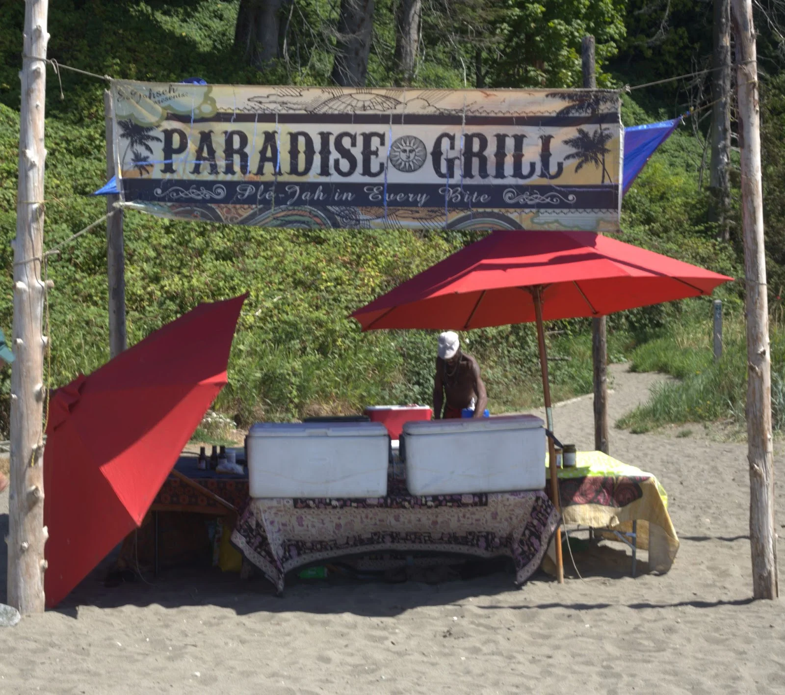 I make for a large banner that says Paradise Grill. Underneath it, a man in khaki cargo pants is carrying coolers and cooking supplies into the shade.