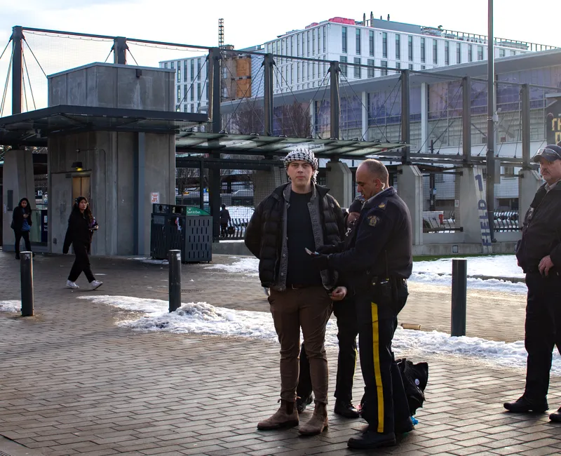 A man staring into a camera on the sidewalk, standing handcuffed by RCMP officers. He is wearing brown pants, a black jacket and a keffiyeh. Two police officers search or hold him as a third looks on.