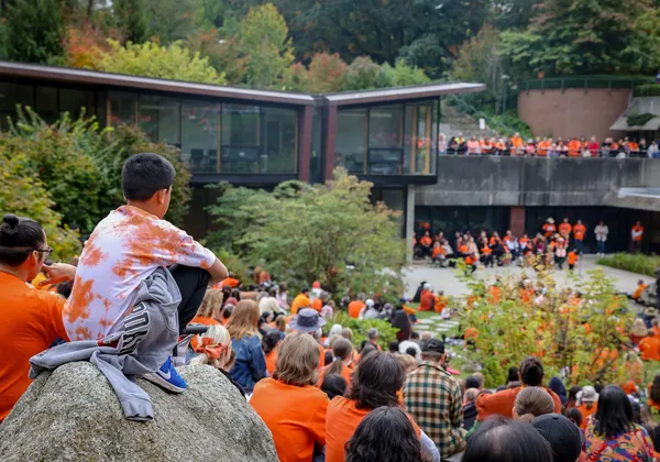 A crowd of people wearing orange shirts are sat in front of the Indian Residential School History and Dialogue Centre.