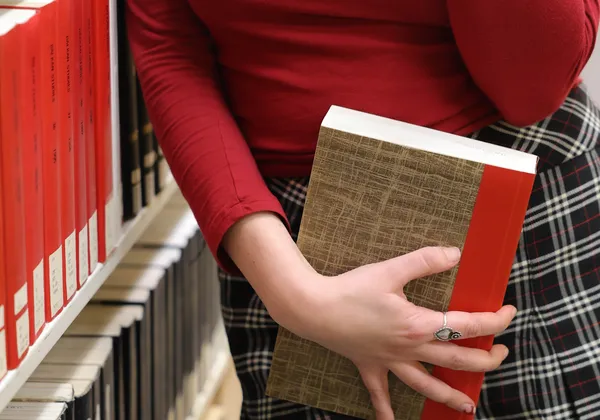 A person in a red top and plaid skirt holds a book over their hips.
