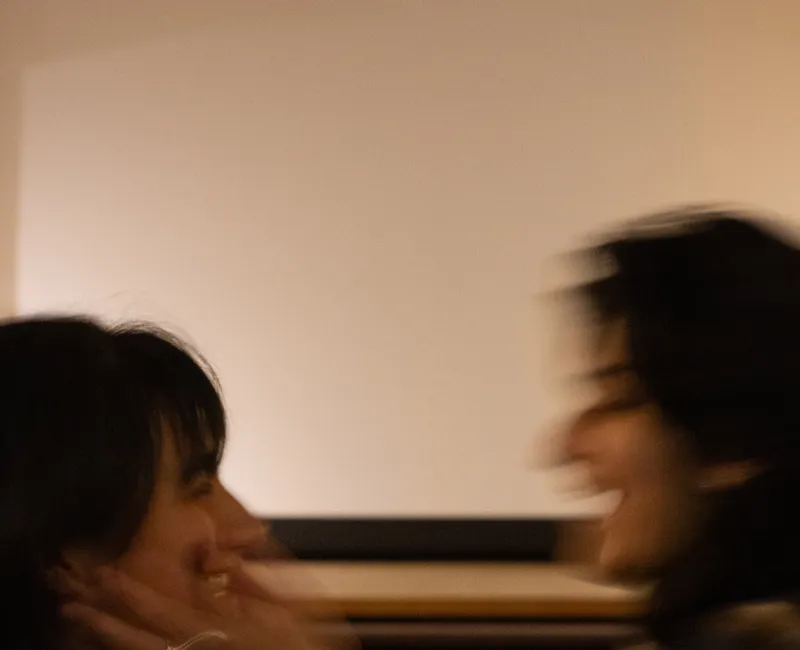 Two girls smiling and sitting in front of a movie screen, one of them cupping the other's face with her hands. Their faces are slightly blurred from movement.