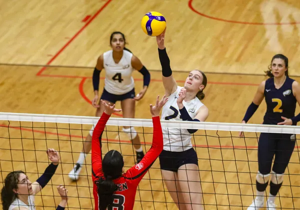 A female volleyball player in a white jersey and black shorts reaches over the net to hit the yellow volleyball. In front of her, another player in a long red sleeve jersey starts to jump up to block the ball. There is another player in white jersey behind her and all players are on a hardwood, indoor court.