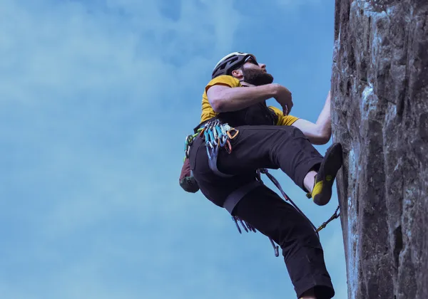 A bottom-up perspective image of a man in a yellow shirt, blue pants and a white bike helmet climbing up a rock face. He is wearing a hip harness with carabiners and a chalk bag attached. He is off-center, with the clear blue sky to the left of the image and the rock face to the right.