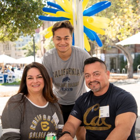 Color photo of three people in Cal t-shirts, two sitting and one standing, all smiling at the camera with blue and gold balloons in the air behind them