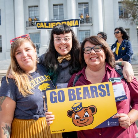 Color photo of three people of varying ages, smiling into the camera, wearing Cal gear and holding a sign that reads