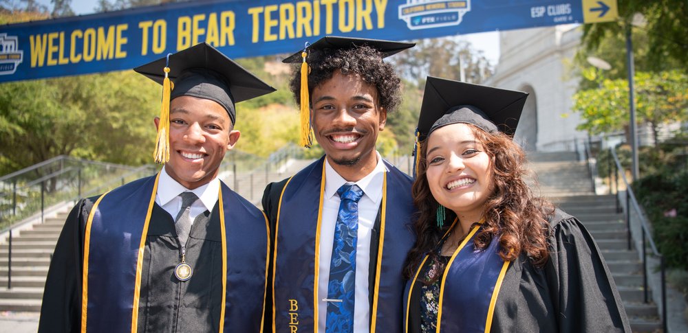 Two male graduates and one female graduate, all in regalia, stand smiling in front of the steps leading to Memorial Stadium.