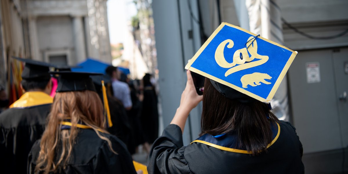 Graduates line up behind the stage at the Greek Theatre to prepare to walk. The woman at the front wears a cap decorated with the Cal script and a bear.