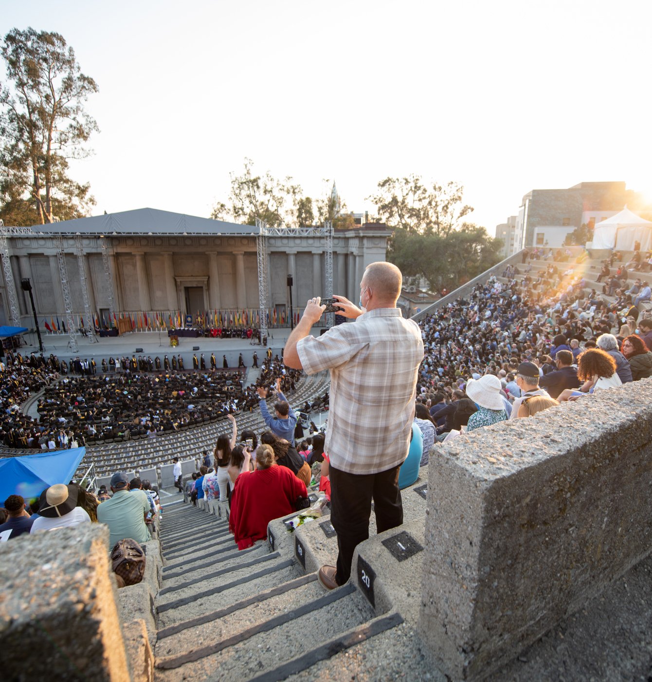 A man in a plaid shirt is standing at the back of the Greek Theatre filming the ceremony on stage.