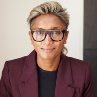 person with short blond hair, glasses, and a burgundy blazer gazes at camera