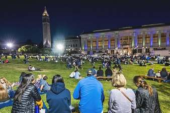 Audience sitting on Memorial Glade with the Campanile and Doe Library lit up blue and gold.