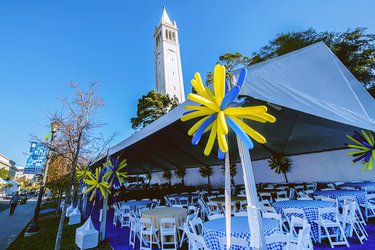 Color photo of BBQ seating in a tent with blue and gold balloon in front of the Campanile