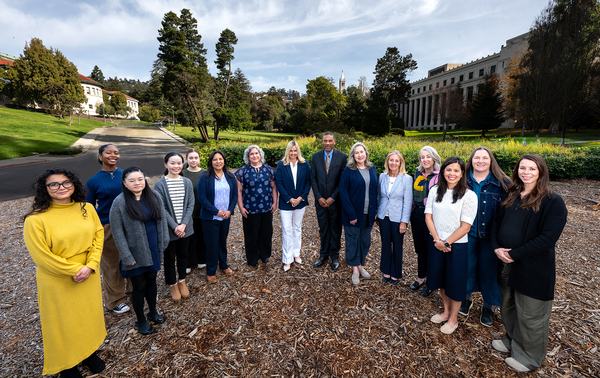 Color photo of 15 people, standing in a semicircle on campus and smiling at the camera.