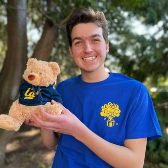 Color photo of Thanasis in front of trees, in a blue shirt, smiling while holding a bear plushie.