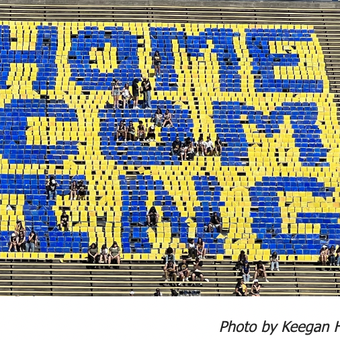 Photo of student in football stands spelling out a "homecoming" with blue and yellow papers.