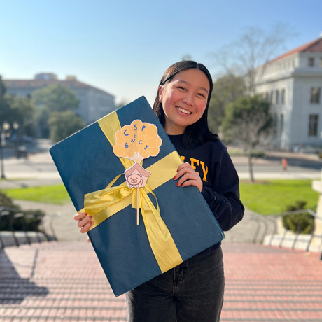 Color photo of Rena smiling & holding a large gift box. Bricks and grass in background.