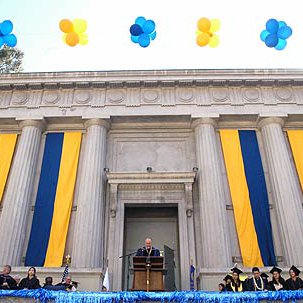 Image of Greek Theatre, with balloons up top and banners in blue and yellow coming down the columns. There are dozens of staff and graduates on stage in their regalia preparing for the ceremony. The sun shines brightly behind the theatre.