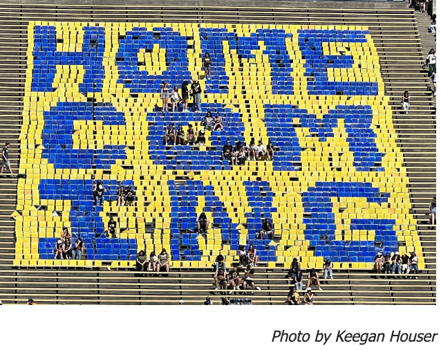 Photo of student in football stands spelling out a "homecoming" with blue and yellow papers.