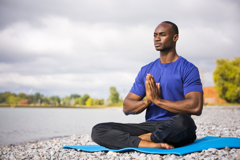  A shirtless man with black pants is meditating on a blue mat on a beach with a lake in the background.