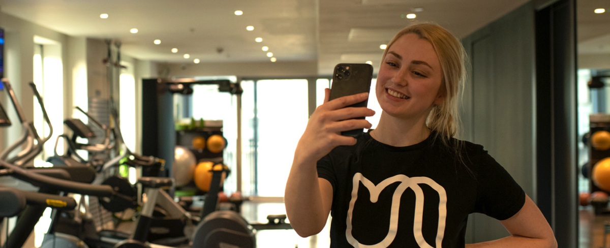 A smiling urban therapist is holding up the phone for a video call. She is standing with her back to a mirror in a gym. 