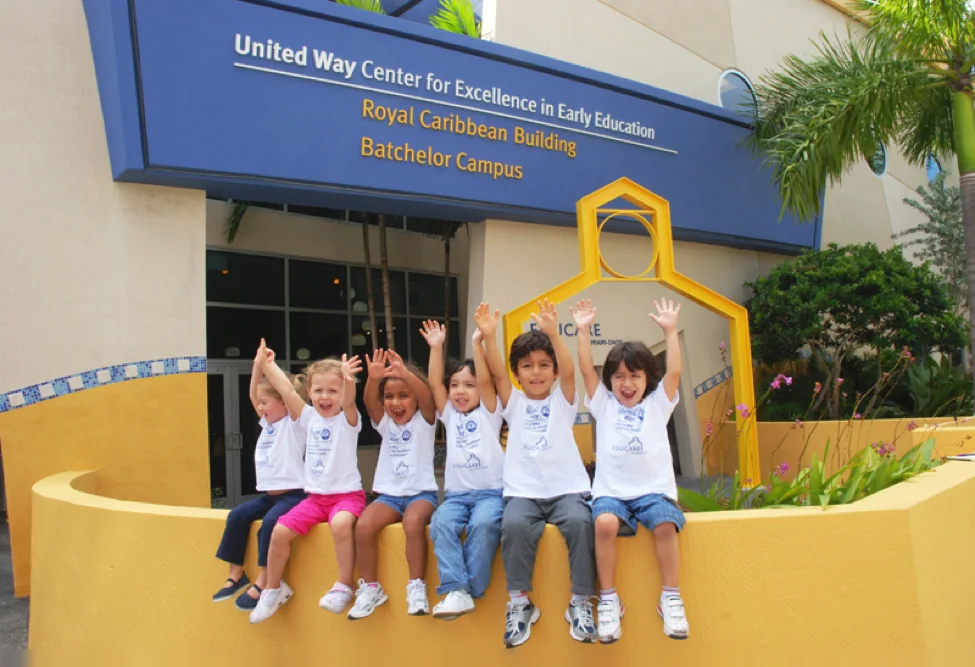 Children Raising their hands and smiling in front of the United Way Center for excellence in Early Education