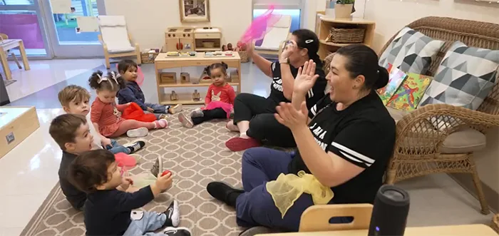 A classroom, you see young children and teachers sitting and singing