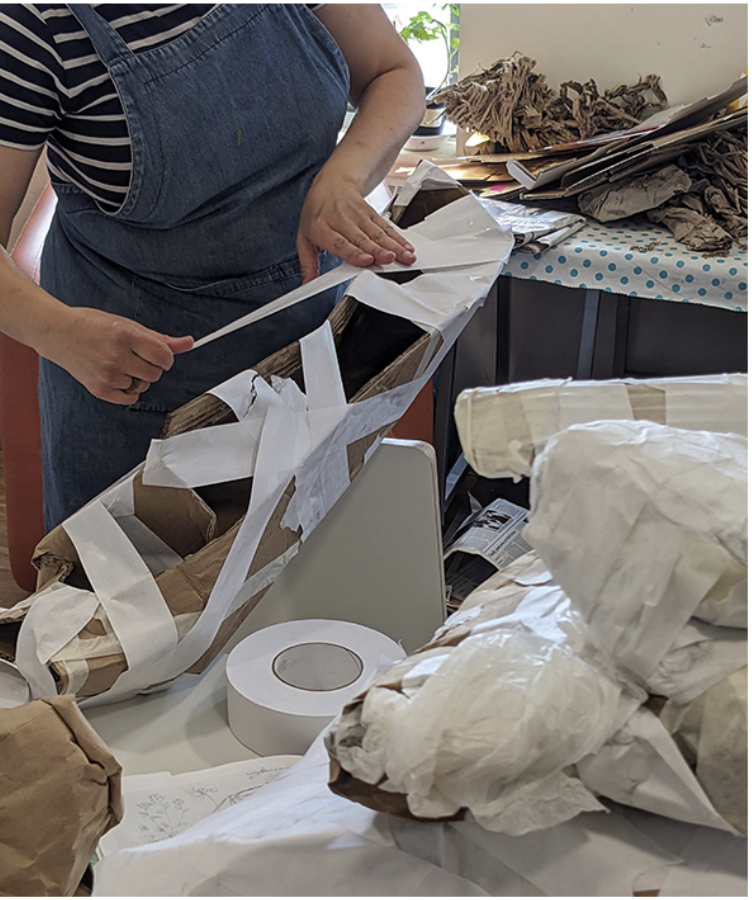 A close of a person wrapping a cardboard shape with lots of strips of white gum tape. There are other cardboard shapes nearby that have already been wrapped