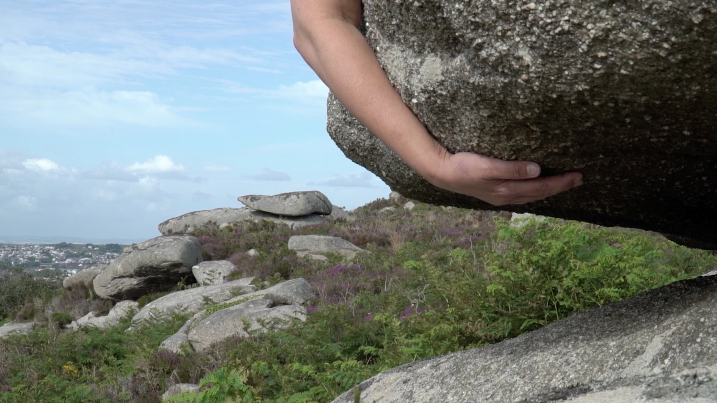 A photograph of a Cornish landscape with a large boulder which has an arm wrapped around it