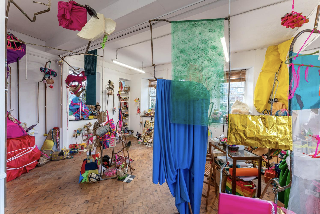 Various colourful objects hang from the ceiling, are placed on a parquet floor and against different temporary structures