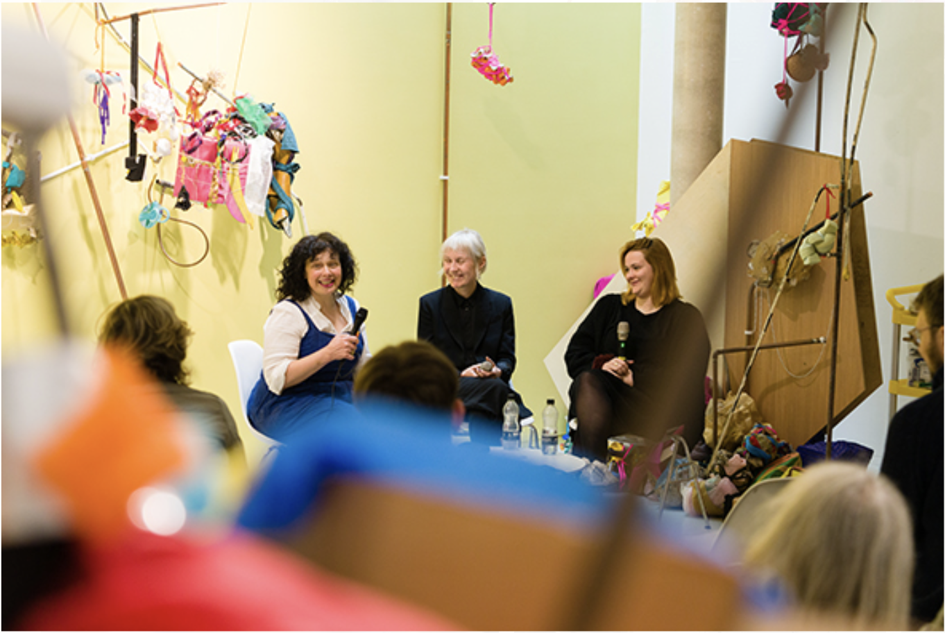 Three women sitting in a room surrounded by colourful objects and shapes.  They are speaking to a seated crowd and smiling