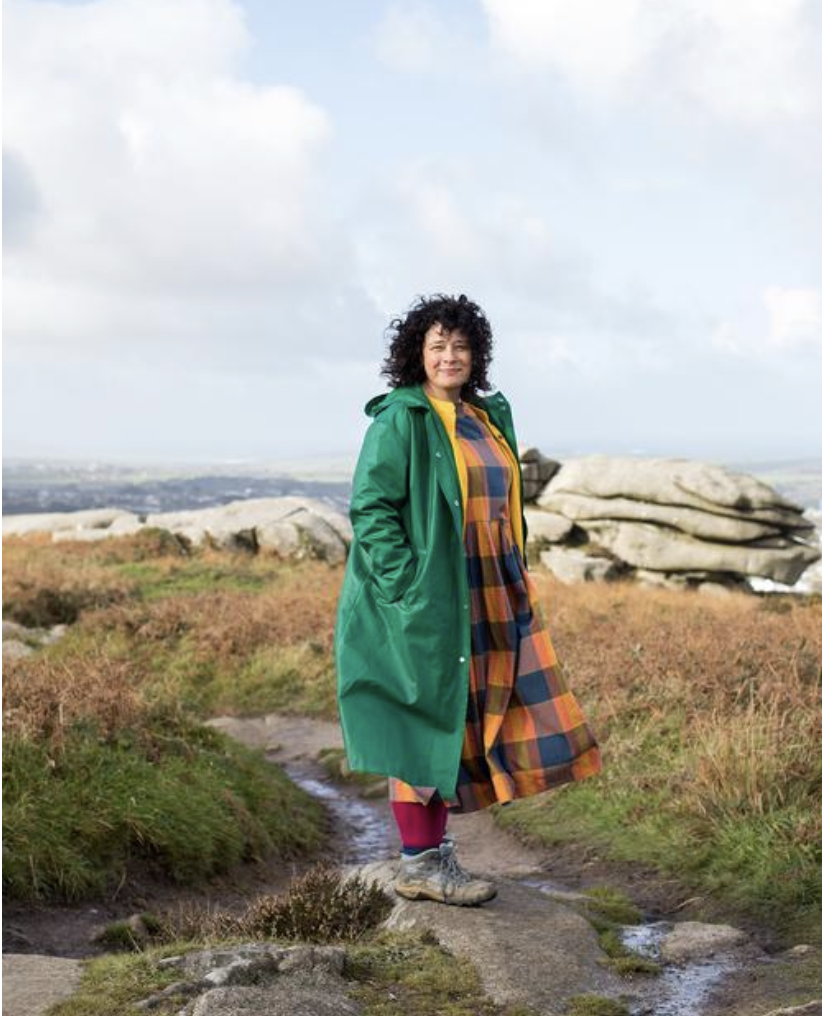 A women in green coat and plaid dress stands smiling for the camera on a rocky hill, surrounded by nature.