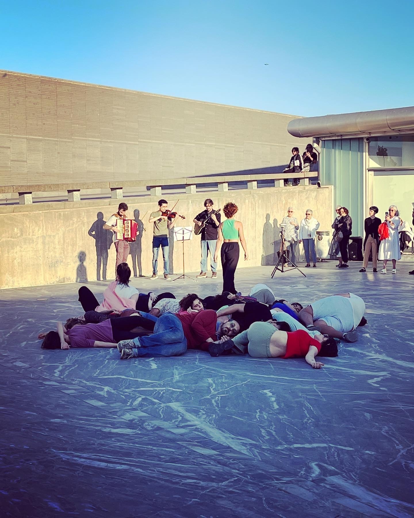 An open public space in front of a building, where a number of people are lying on the ground together.