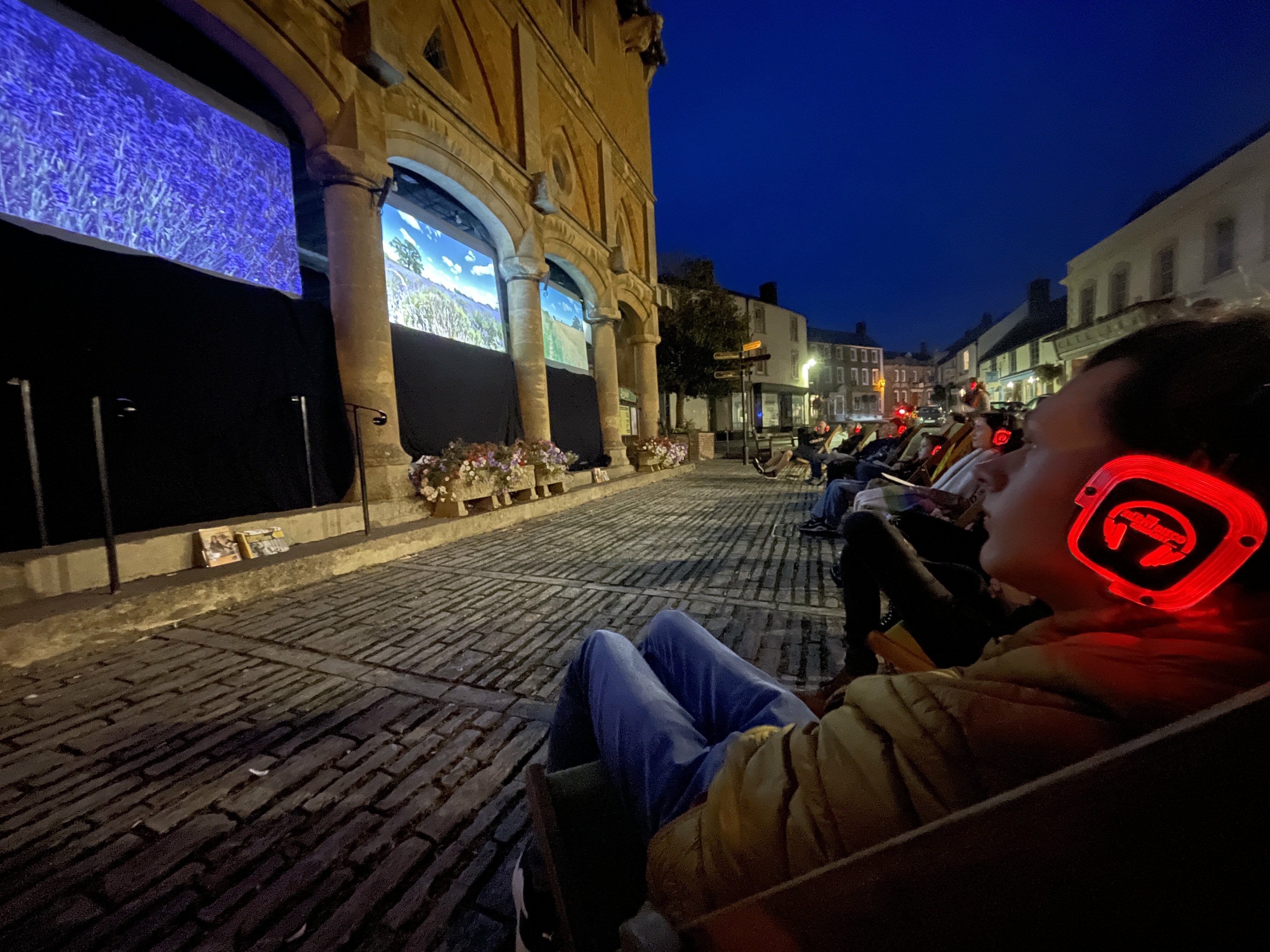A group of people sitting in a public space watching a multi screen film installation projected on the side of a historical building.