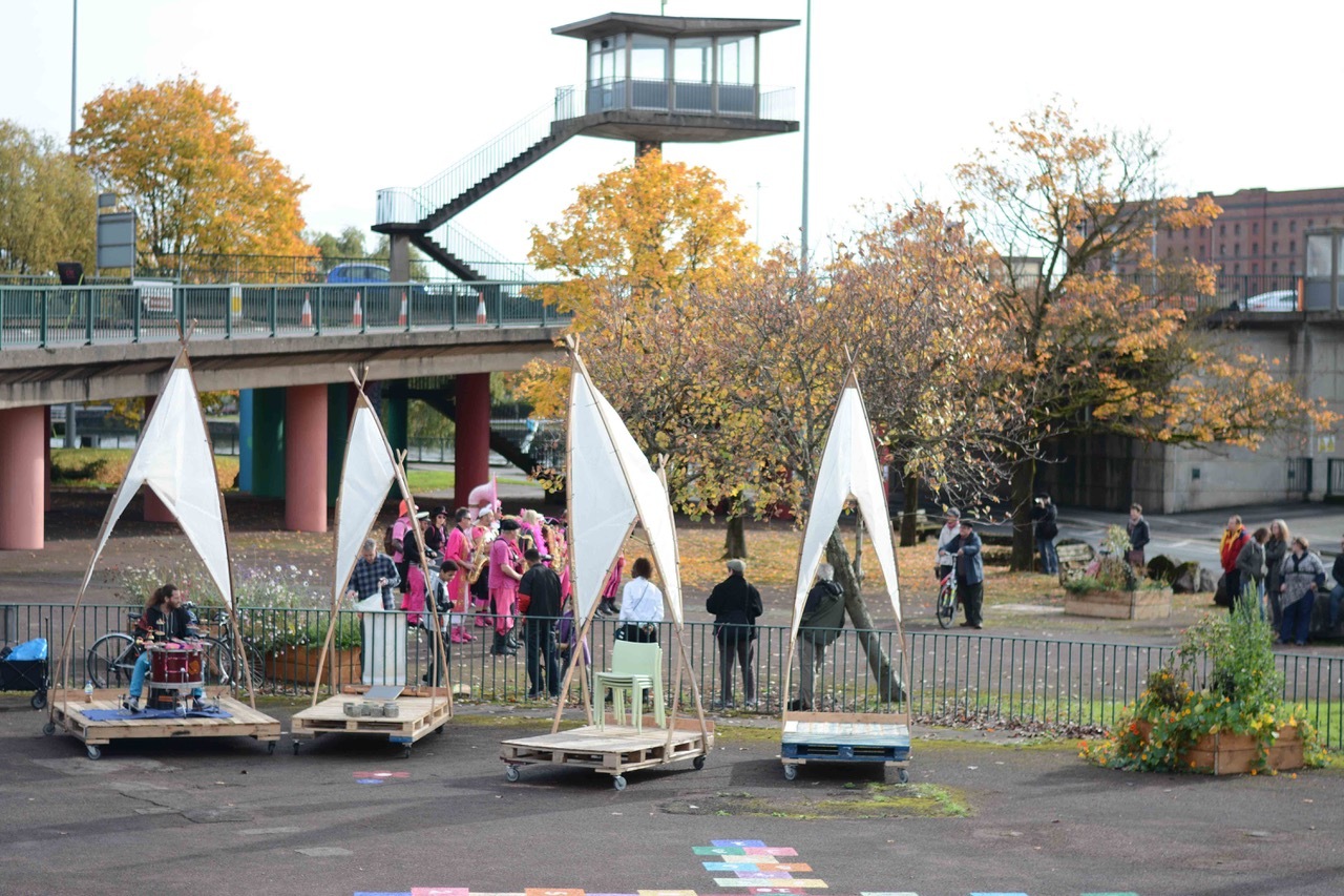 People gathered in a large area under a flyover, next to four sculptures constructed of wooden platforms, timber poles and white fabric.