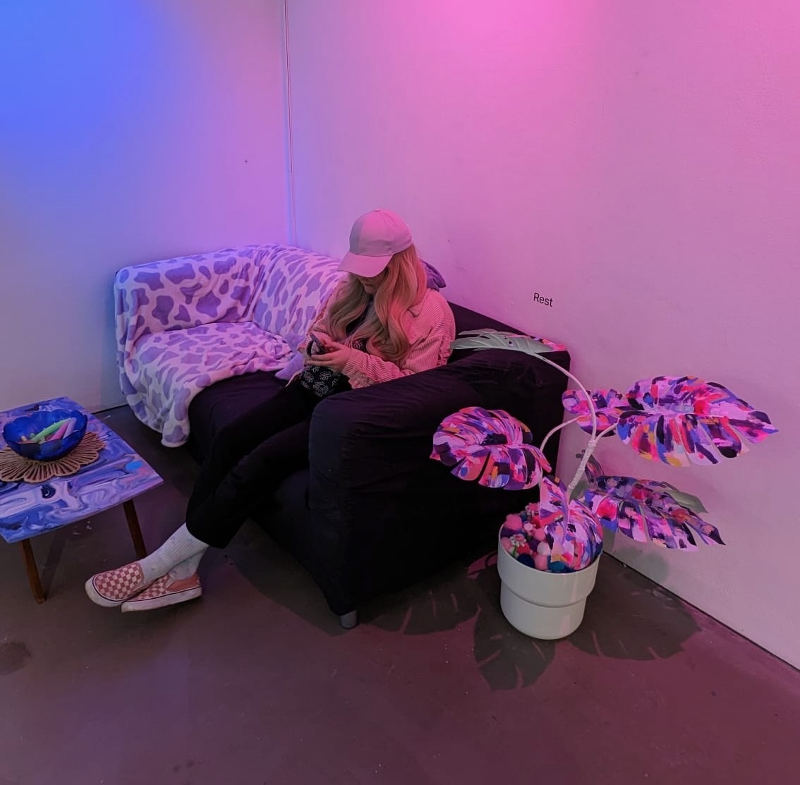 A person with long hair wearing a cap, sitting on a sofa is a softly purple-lit room, next to a colourful pot plant and low table