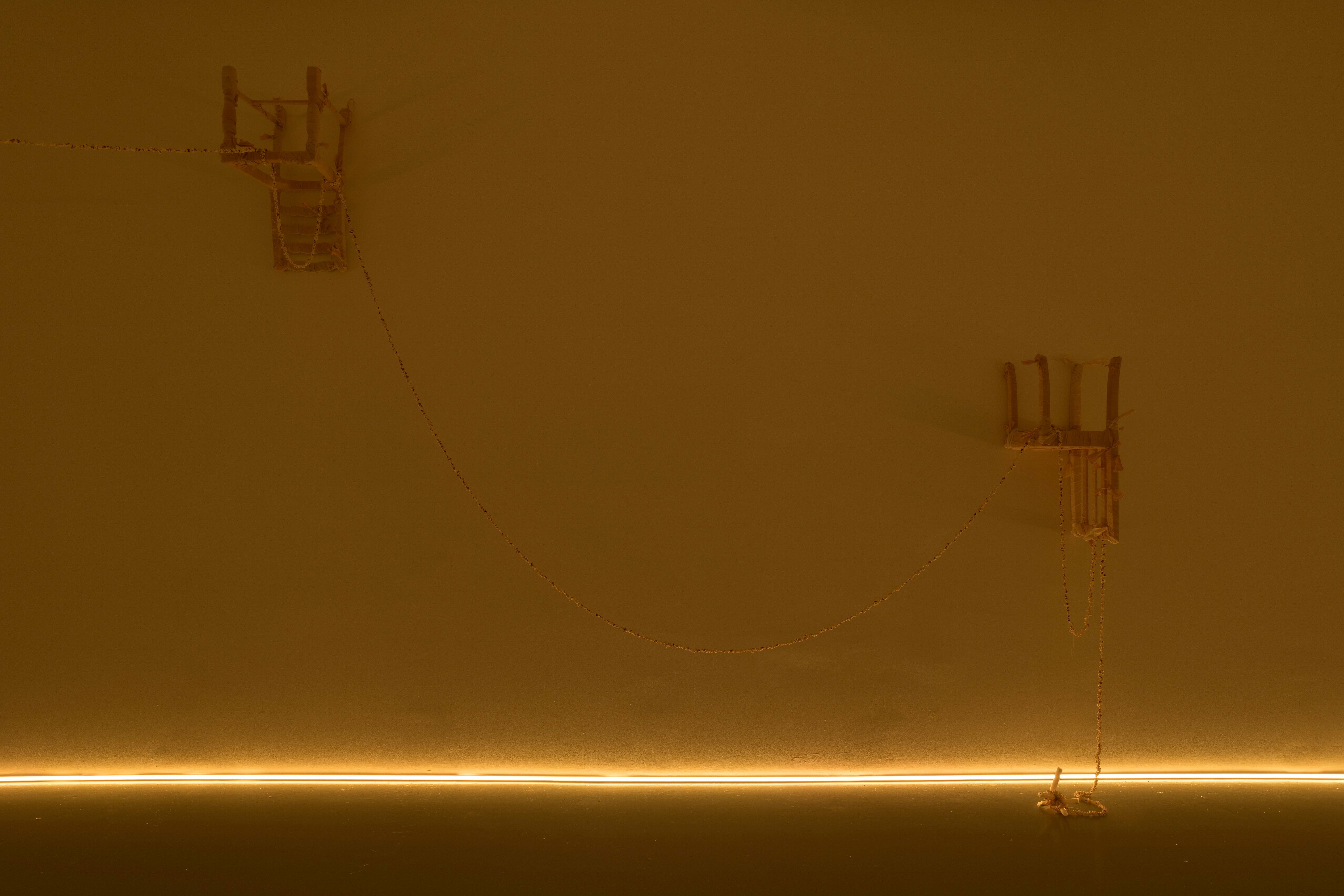 A photograph of part of an exhibition installation, consisting of two chairs that have been hung on a wall upside down. The chairs are wrapped in white fabric, and are connected by a string of popcorn, that continues to the left of the image and to the right where it hangs from one of the chairs and pools on the floor, wrapping around a candle. There is a strip of yellow lighting where the wall meets the floor, creating a low warm light.