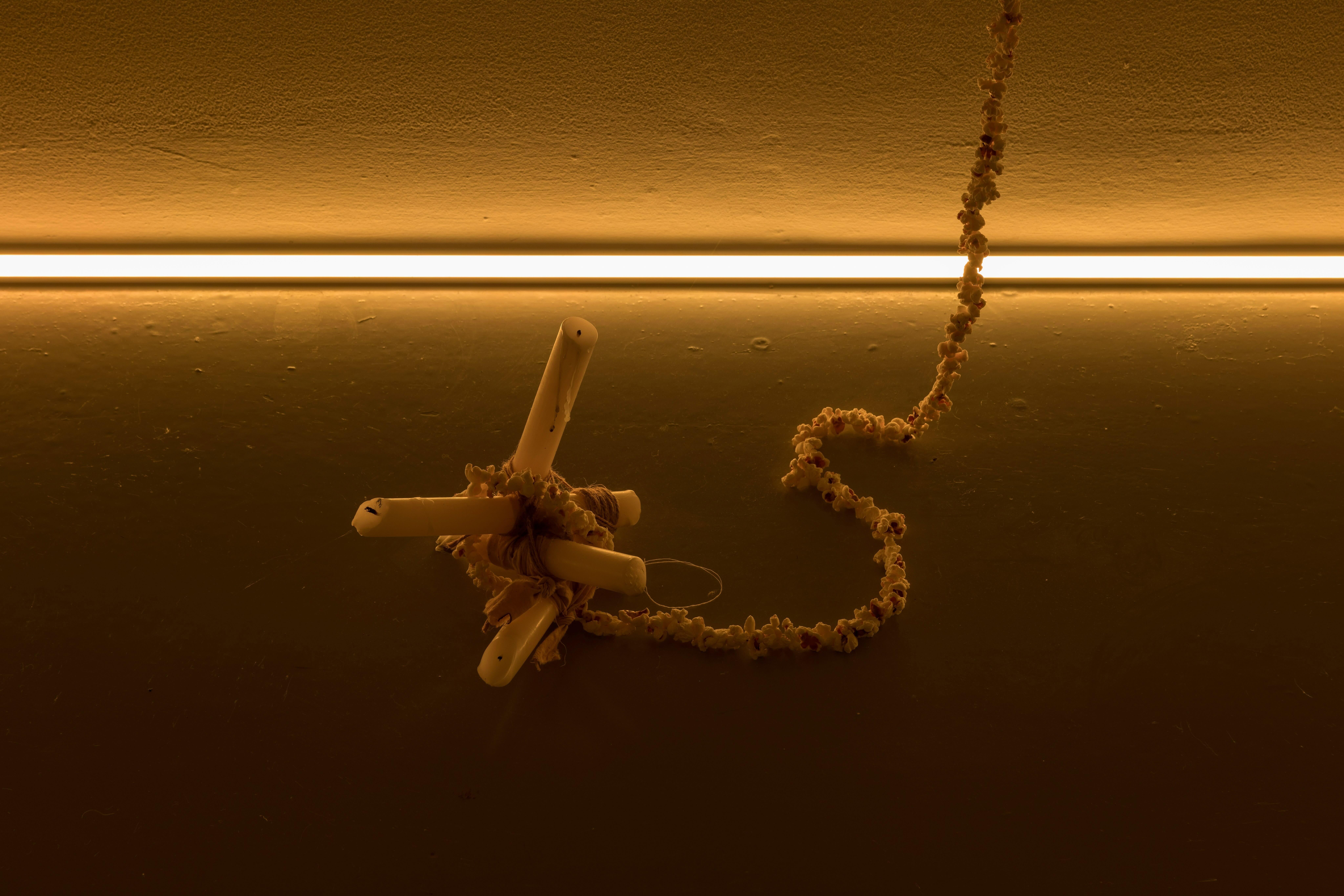 A photograph of an artwork in a gallery space, consisting of a small sculpture places on the floor made from four candles which are joined together by a string of popcorn, which continues along the floor and up towards the ceiling. There is a strip of yellow lighting where the wall meet the floor, creating a low, warm light.