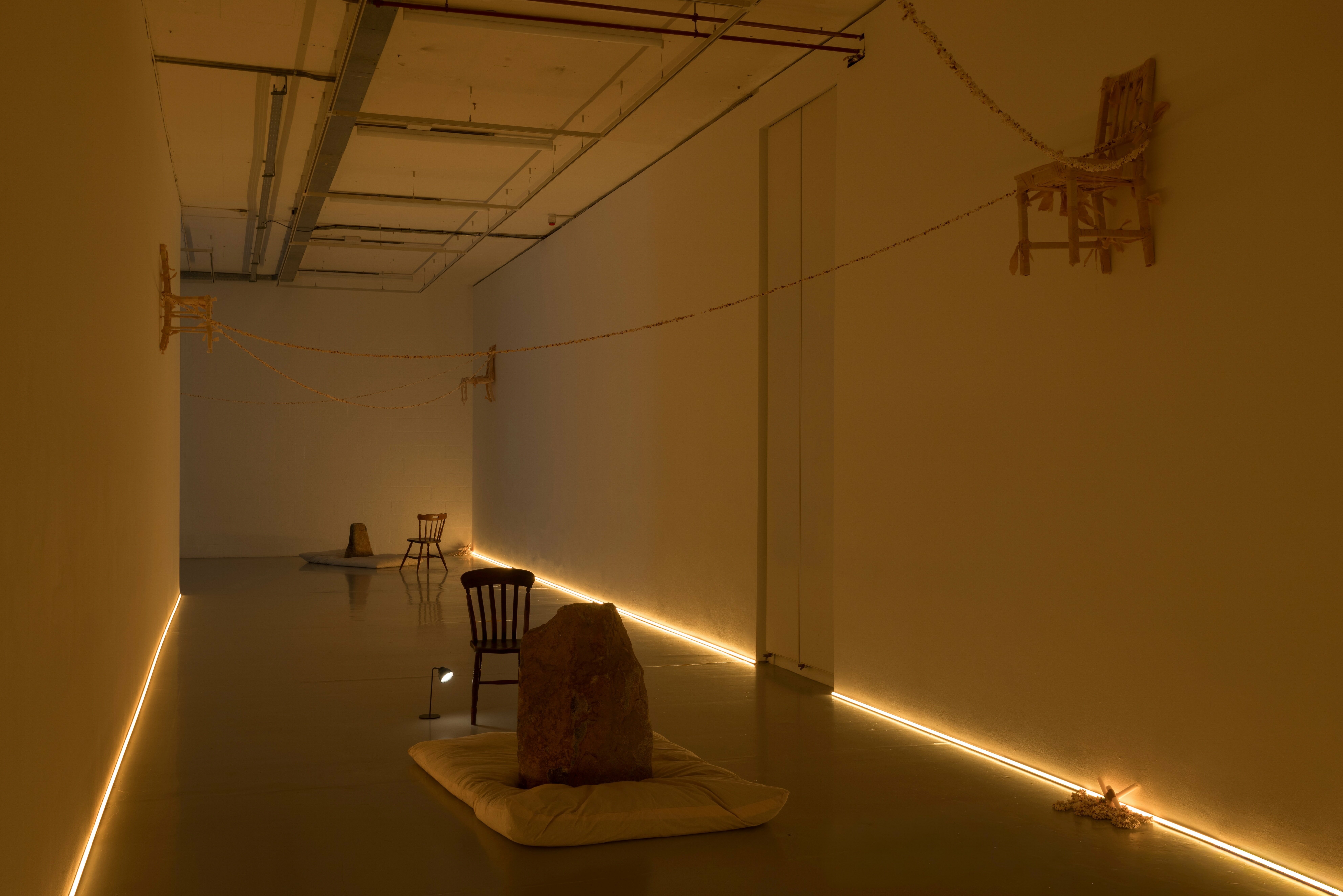 A photograph of an exhibition installation in a large gallery space, consisting of large rocks positioned on rectangular off-white floor cushions. There are chairs and small lamps close to the cushions, and three chairs that have been wrapped in fabric are hung on the far and right walls and on the ceiling. Strings of popcorn connect the chairs and are wrapped around one of the central pillars in the space. There is a strip of yellow lighting where the walls meet the floor, creating a low, warm light throughout space.