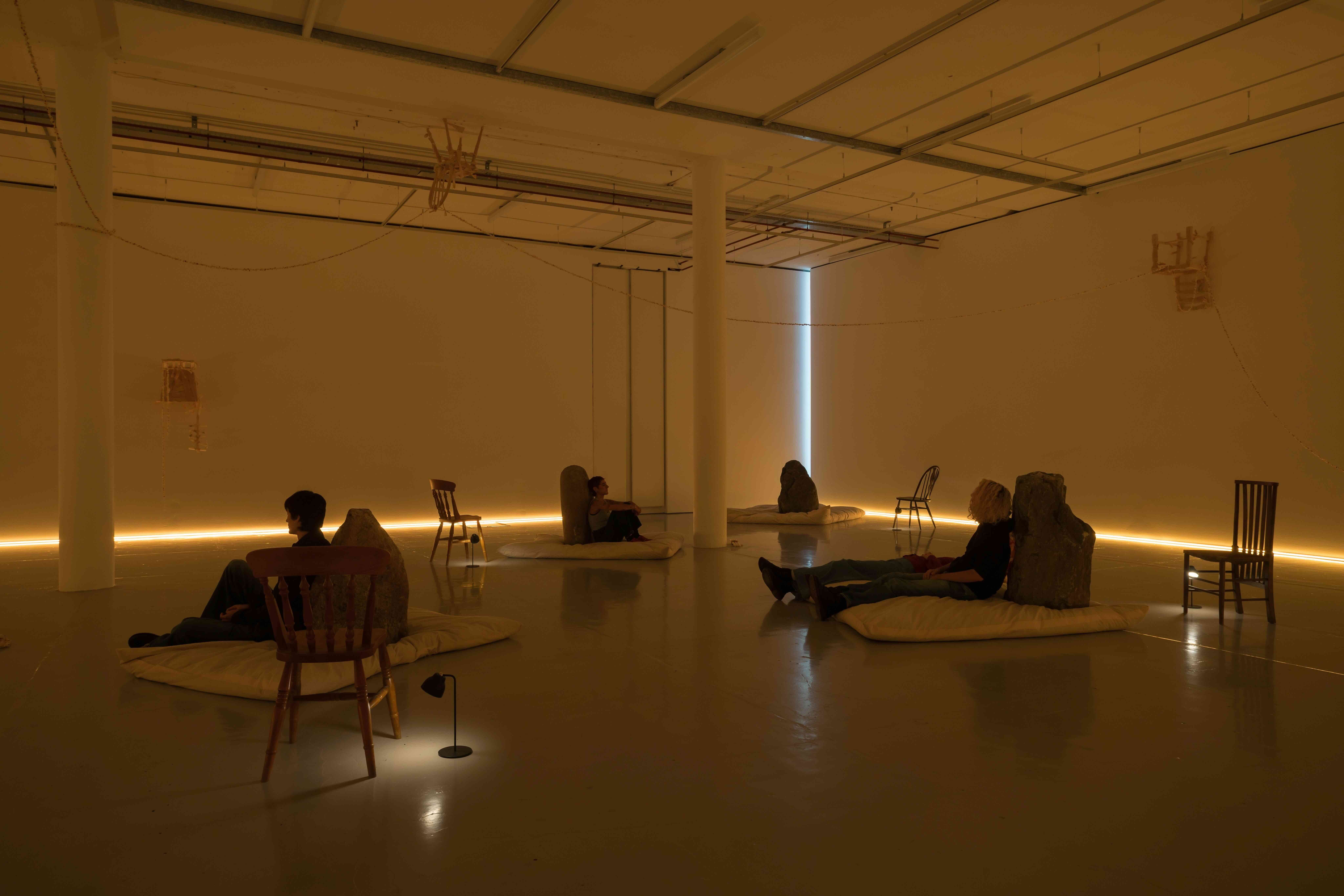 A photograph of an exhibition installation in a large gallery space, consisting of large rocks positioned on rectangular off-white floor cushions. There are chairs and small lamps close to the cushions, and three chairs that have been wrapped in fabric are hung on the far and right walls and on the ceiling. Strings of popcorn connect the chairs and are wrapped around one of the central pillars in the space. There is a strip of yellow lighting where the walls meet the floor, creating a low, warm light throughout space. Three people are sitting on the floor cushions, leaning against the rocks.