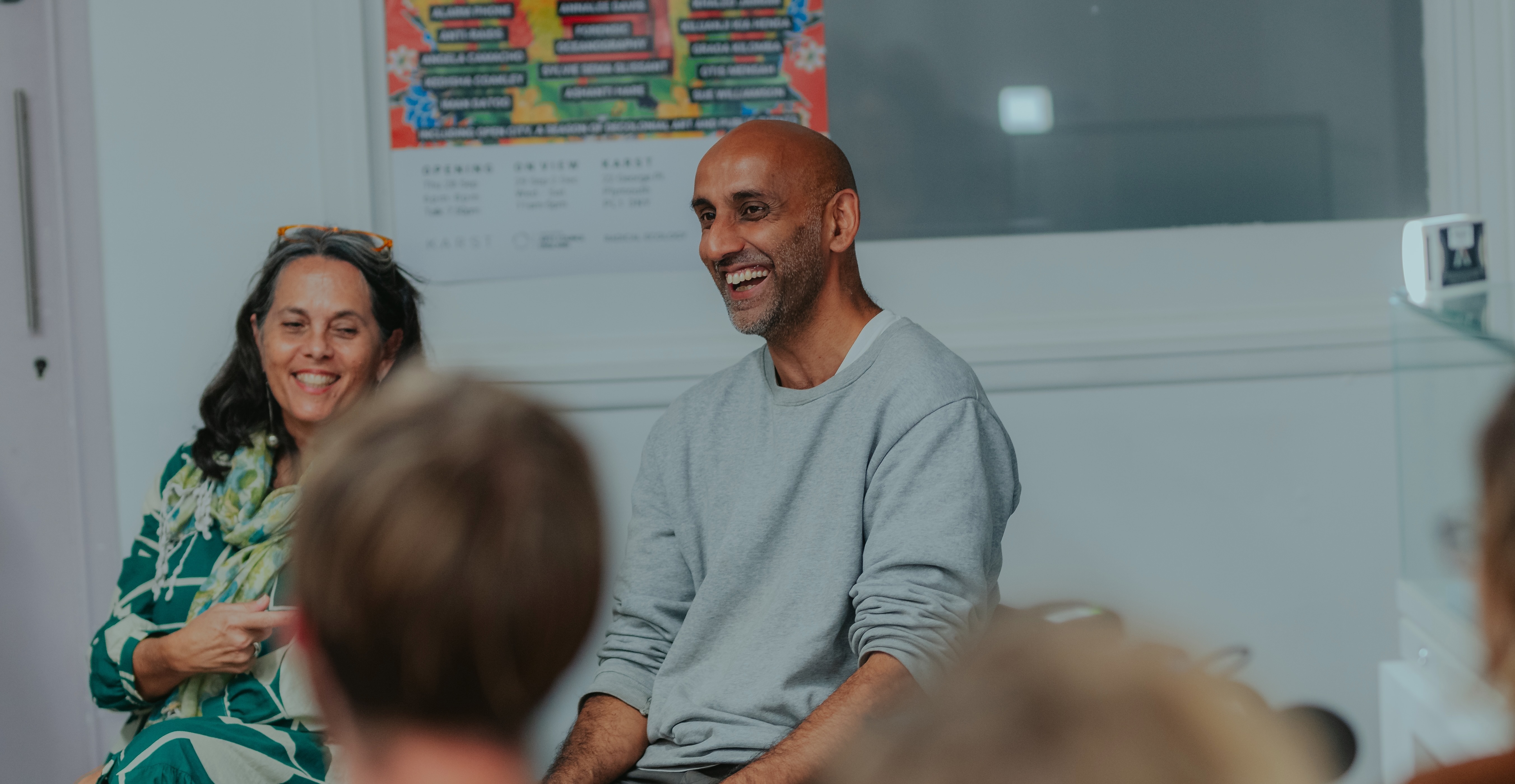 Two people sitting in front of an audience in a brightly lit room. The woman is wearing a green dress and the man has on a light grey sweatshirt. Both are laughing,