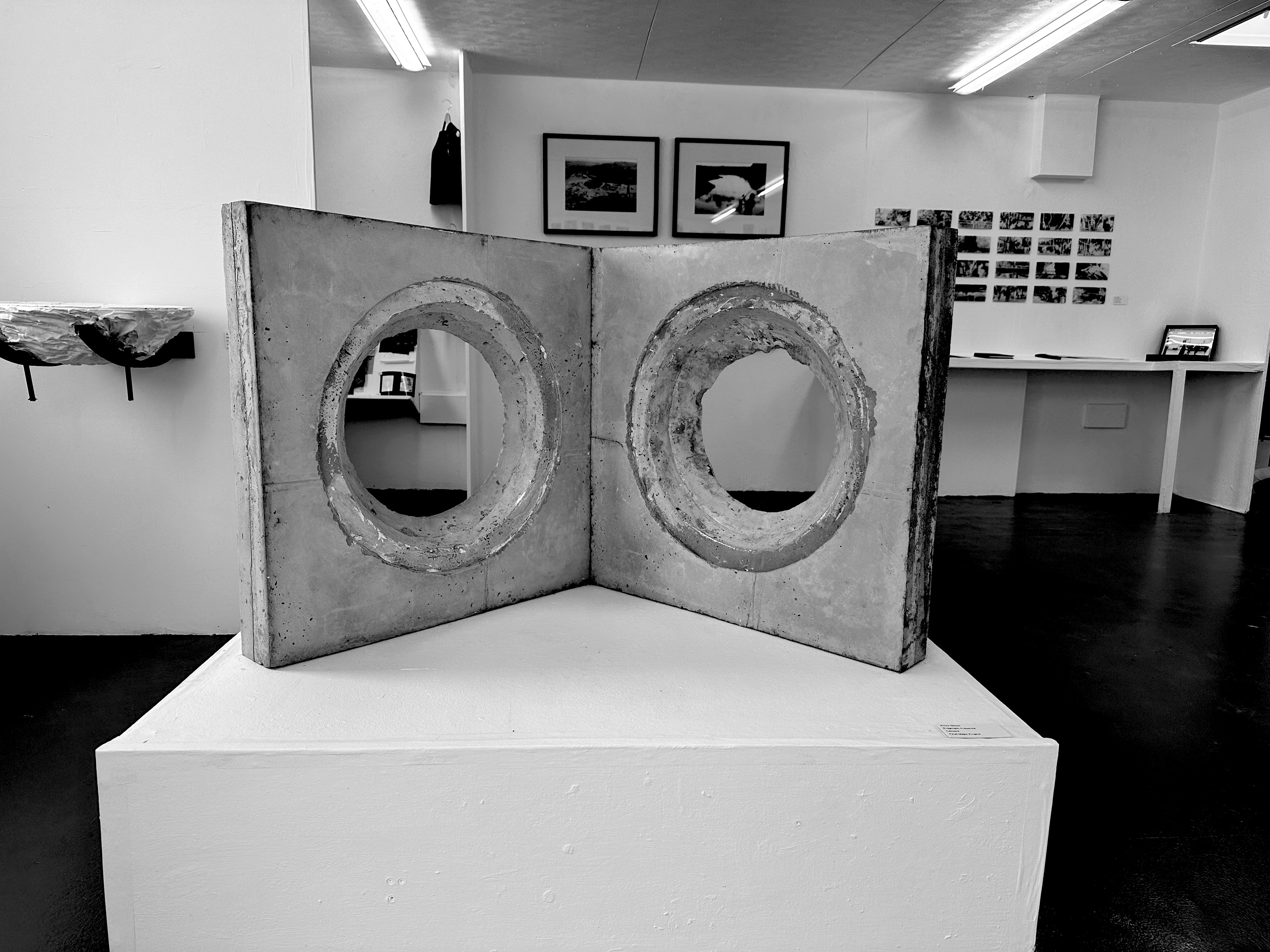 A stone sculpture is sat on a large white plinth. The sculpture is two large concrete squares joined at the centre, stood upright and open like a book. There are two large holes punctured through the middle of both slabs