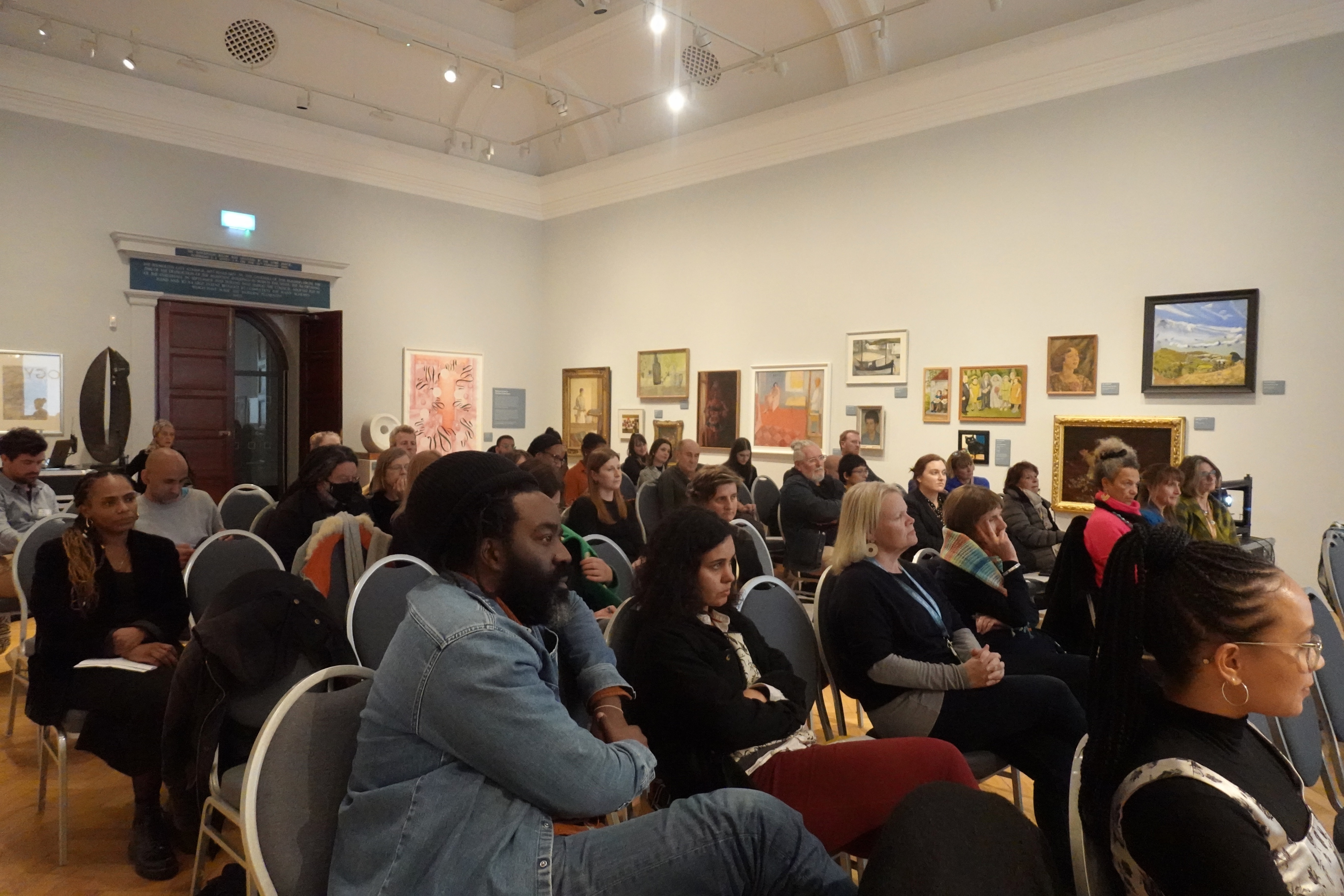 A room full of people sat facing forwards, they are in a museum with paintings hung on the wall in a salon formation.