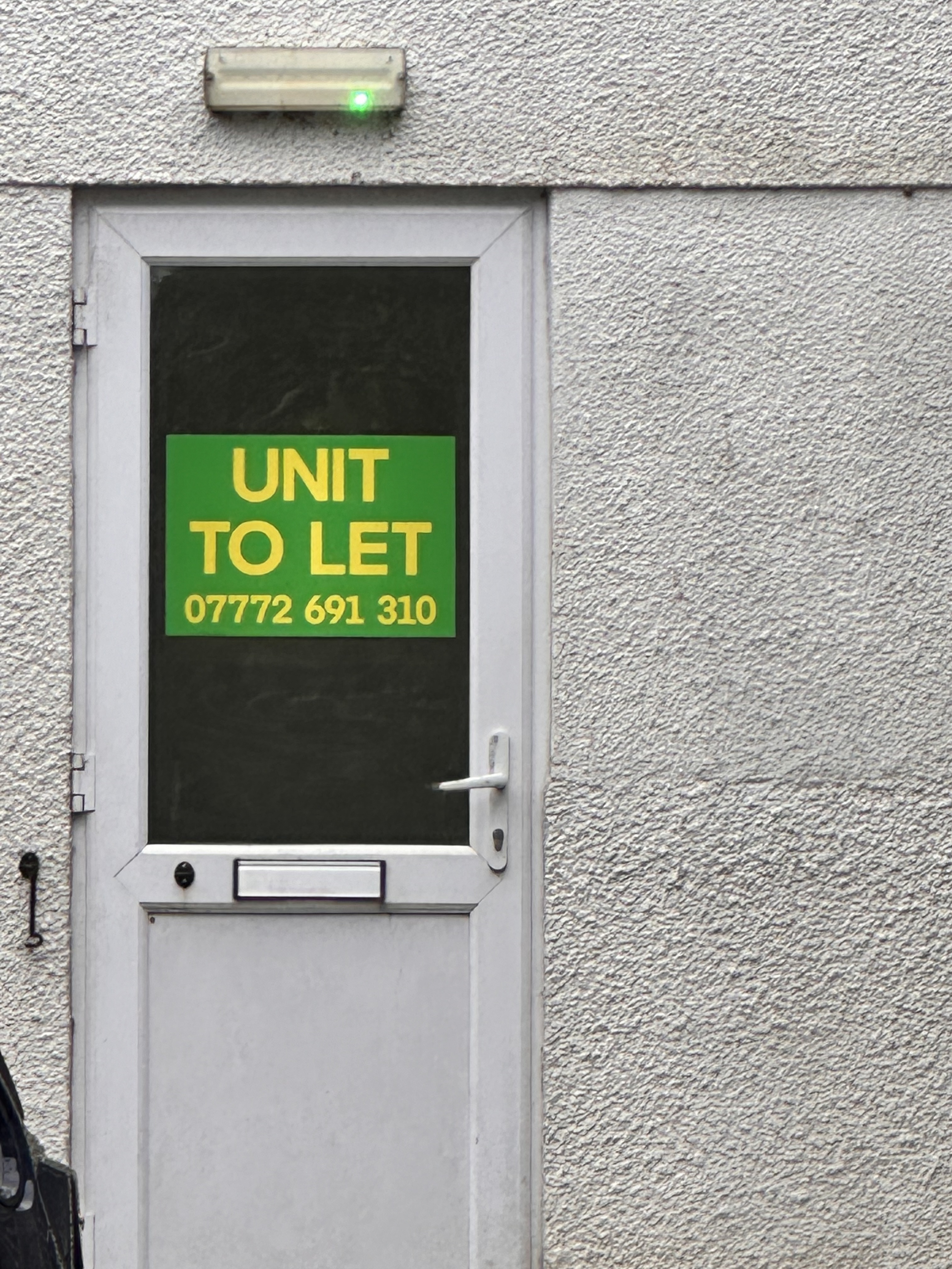 A white framed door mounted on a white rendered building has a sign stuck to the glass panel that says 'UNIT TO LET' and a phone number to call.