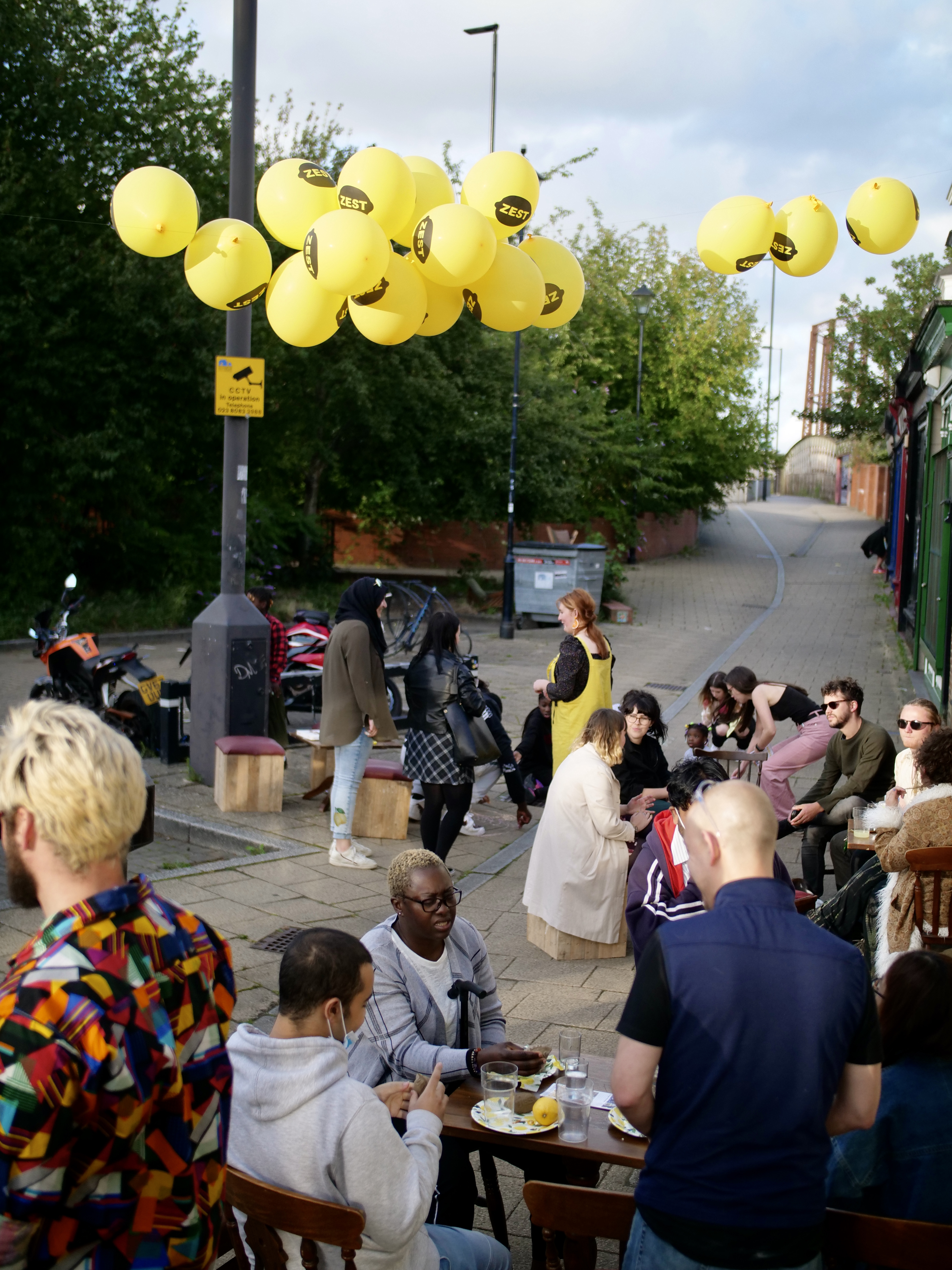 Groups of people are sat outside at cafe tables on a pedestrian street, there are yellow balloons above with the word ZEST printed on them