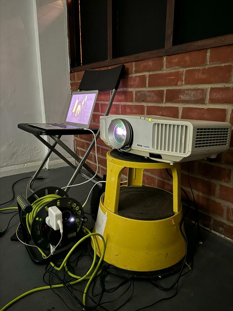 A projector placed on top of a yellow step stall plugged into an extension cable reel and a laptop placed on a chair