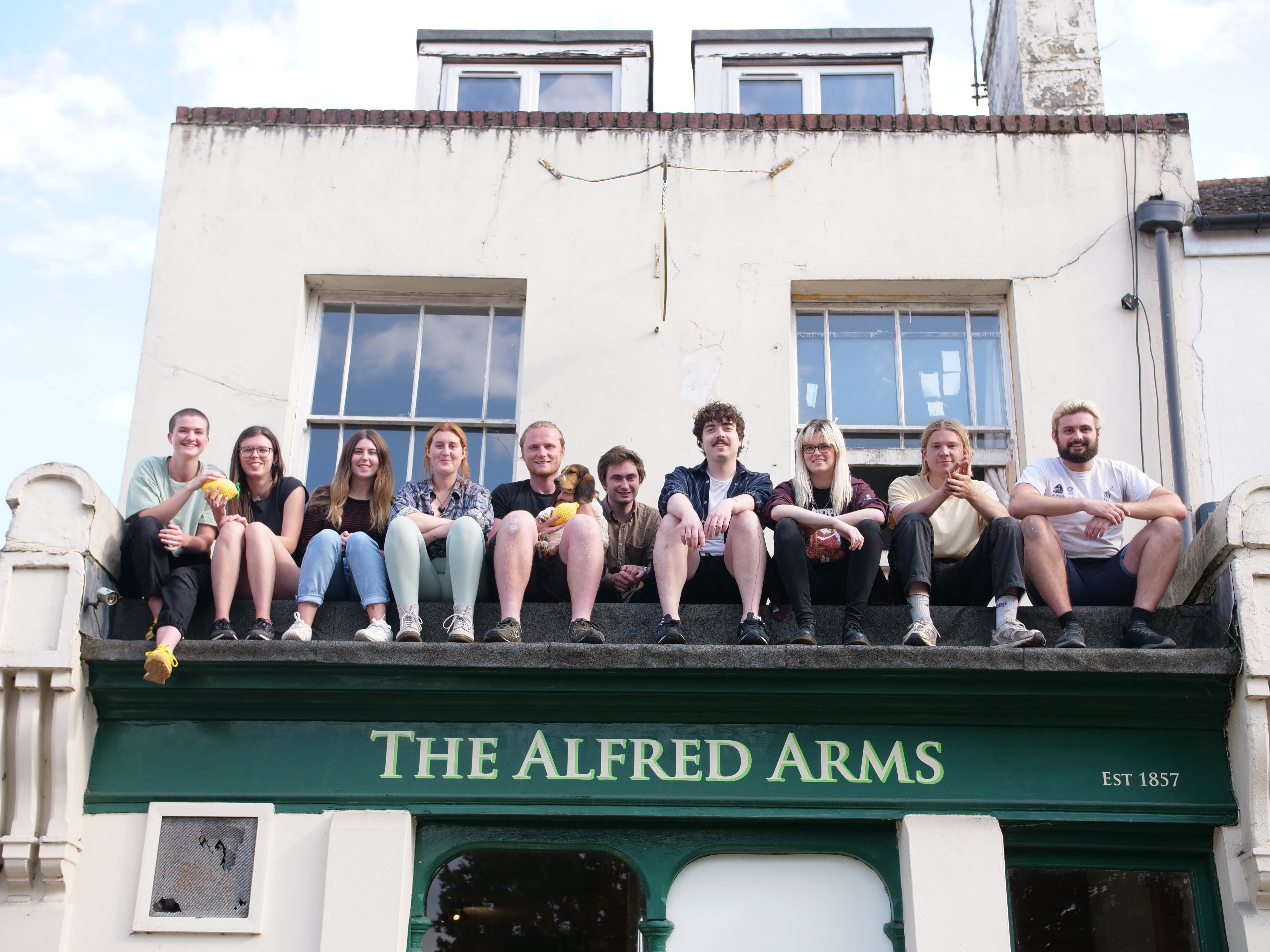 A group of people are sat in a line on a flat roof of a pub called The Alfred Arms, smiling and looking down for the camera