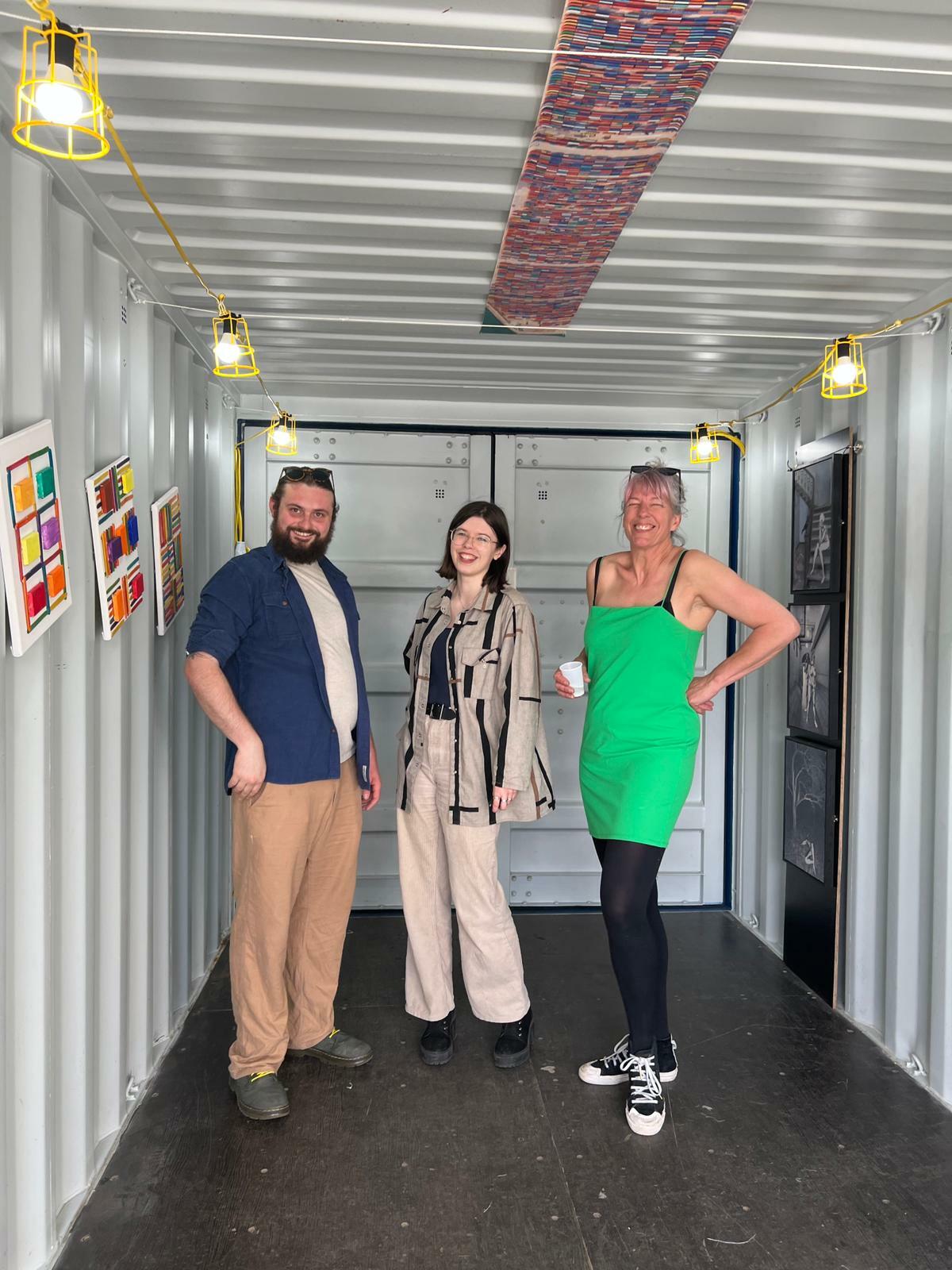 An image of three people stood inside a shipping container smiling for the camera. there are artworks hanging on the walls and ceiling and a string of yellow spotlights along the walls