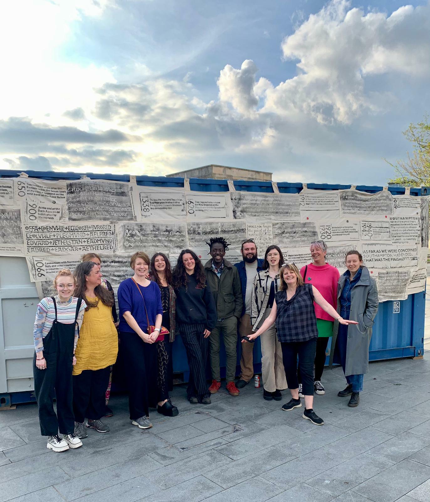 A group photograph of people smiling for the camera, stood in front of a shipping container that has printed fabric hung on its side