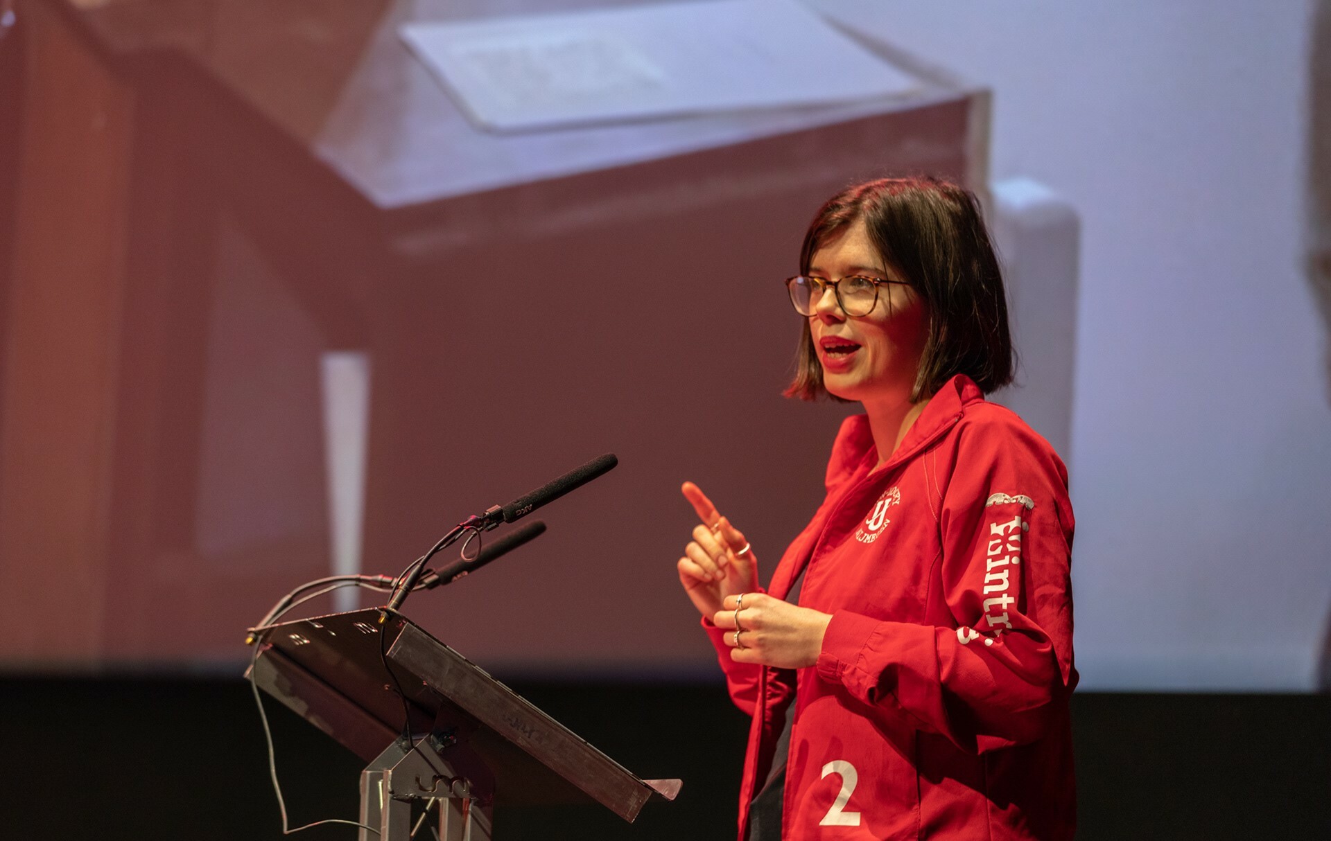 A woman in a red jacket giving a presentation stood at a lectern.