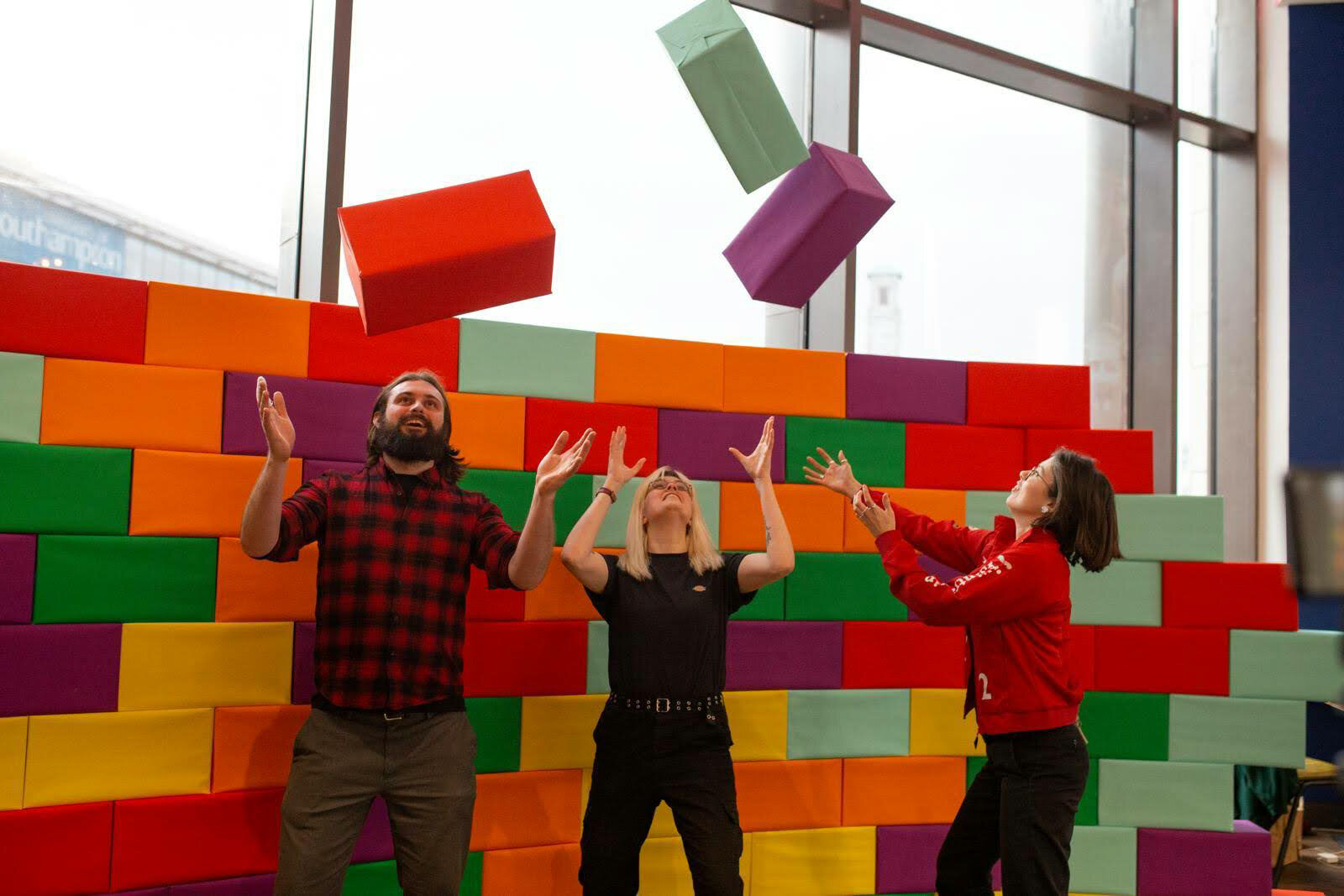 Three people are smiling, throwing three colourful block shapes into the air , behind them is a colourful block wall made of of the same shapes