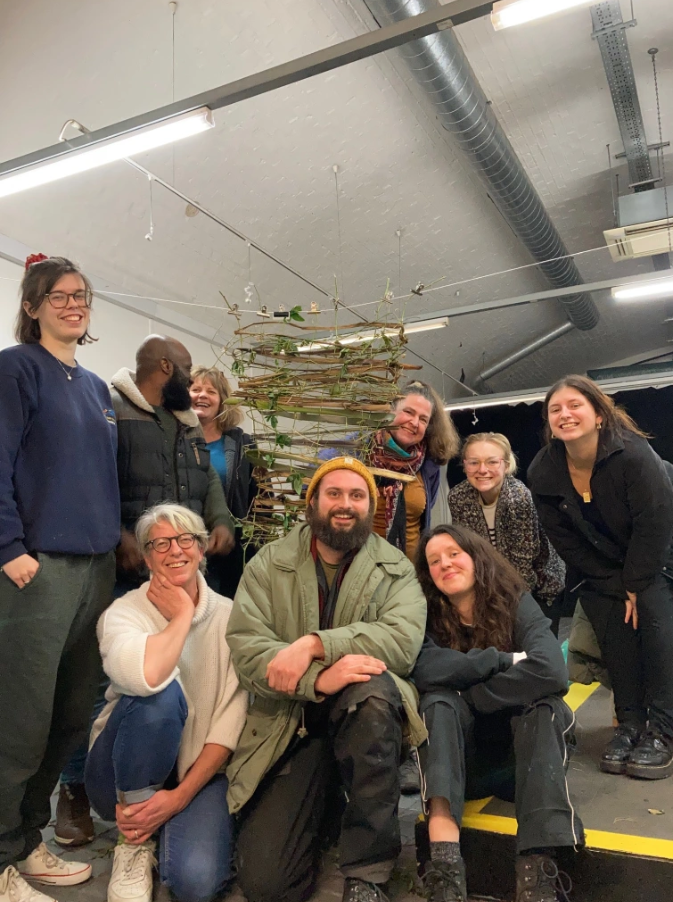 A photograph of a mixed group of people stood and crouching, smiling for the camera, in the middle of the group is a hanging sculpture made from thin strips of wood and greenery